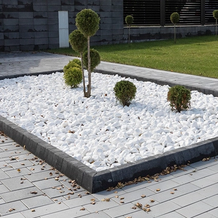 Kostka brukowa i kamień ozdobny - Kwadratowa rabata ogrodowa otoczona ciemnym, betonowym krawężnikiem, wypełniona białym kamieniem ozdobnym i niskimi, formowanymi krzewami. W tle widoczna szara kostka brukowa.