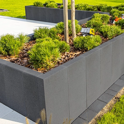 Kostka brukowa i kamień ozdobny - Podwyższona rabata w ogrodzie, zbudowana z ciemnych, betonowych palisad, wypełniona korą i zielonymi krzewami. W tle widoczny trawnik i fragment jasnego chodnika.