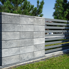 Kostka brukowa i kamień ozdobny - Ogrodzenie nowoczesne, łączące szare bloczki betonowe z metalowymi panelami. W tle widoczne drzewa iglaste i trawnik na pierwszym planie.