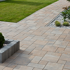 Kostka brukowa i kamień ozdobny - Ścieżka w ogrodzie wykonana z prostokątnej kostki brukowej w odcieniach beżu i szarości, prowadząca do trawnika i rabaty z białym kamieniem.