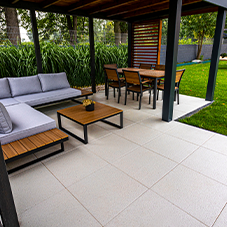 Kostka brukowa i kamień ozdobny - Nowoczesny taras z dużych, jasnych płyt tarasowych. W cieniu pergoli umieszczone są szare meble ogrodowe, stół i narożnik, z widokiem na zielony trawnik.