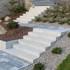 Kostka brukowa i kamień ozdobny - Schody zewnętrzne z jasnego, betonowego kruszywa, prowadzące w górę ogrodu. Ułożone są pomiędzy rabatami ozdobionymi korą, kamykami i kwitnącymi roślinami.